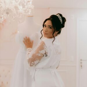 Bride in lace robe with wedding dress in elegant room, showcasing bridal elegance.