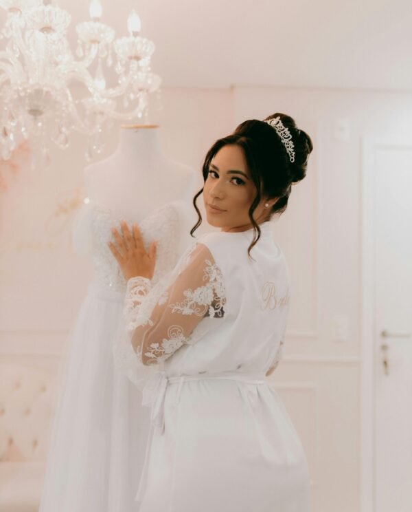 Bride in lace robe with wedding dress in elegant room, showcasing bridal elegance.